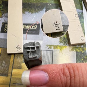 May include: A metal branding iron stamp featuring a raised symbol that resembles a stylized character. The stamp is shown with a circular inset highlighting the impression it leaves on a light-colored rectangular clay or ceramic tile surface.