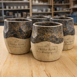 May include: Four handmade ceramic tumblers. The upper section of each tumbler has a speckled brown and tan pattern, with a lighter tan base. The text "Hildur Björk Pottery" is printed on the lower section of each tumbler.