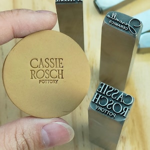 May include: A round, tan pottery piece with the embossed text "CASSIE ROSCH POTTERY" is held near metal stamps. The stamps have reversed text, including "CASSIE ROSCH" and "CERAMICS".