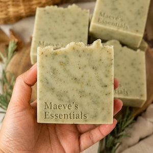Op de afbeelding: Een rechthoekig stuk lichtgroene zeep met zichtbare kruidenstukjes en de tekst Maeve's Essentials in reliëf op de voorzijde. De zeep heeft een getextureerde, onregelmatige bovenrand en een glad, vlak oppervlak met een natuurlijk spikkelpatroon.