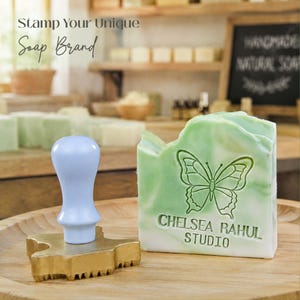 May include: A light blue handled metal stamp with a butterfly and the text CHELSEA RAHUL STUDIO, positioned next to a rectangular bar of green and white marbled soap featuring the same butterfly design and text embossed on its front surface.