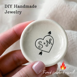 May include: A small, round, off-white ceramic trinket dish with a crackled glaze finish. The center features a black graphic of a heart pierced by an arrow, with the letters S + M inscribed inside. Text on the image reads DIY Handmade Jewelry.