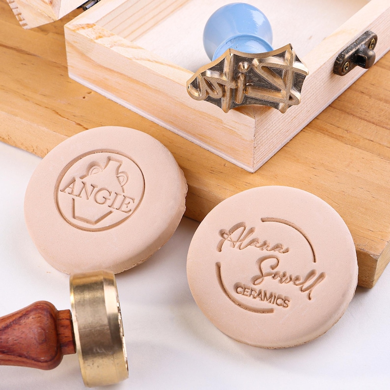 May include: A wooden box with a blue and gold wax stamp and two round, beige wax seals. One seal reads "ANGIE" and the other reads "Alana Sowell CERAMICS". The stamp has a wooden handle and a gold metal stamp head.