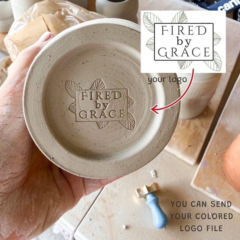 May include: A circular ceramic base featuring a stamped logo that reads FIRED by GRACE, surrounded by a leaf design. The stamp is pressed into the light beige clay surface. A small metal stamp tool with a blue handle is visible nearby on the surface.
