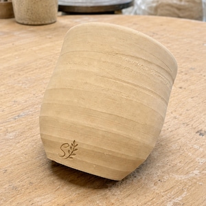May include: A light beige, unglazed ceramic cup with a tapered cylindrical shape and horizontal ridges along the exterior surface. The lower portion of the cup features a small, stamped logo consisting of the letter S next to a leafy sprig design.