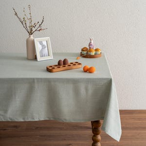 May include: A pale green tablecloth on a wooden table, decorated for Easter. A vase with branches, a framed rabbit print, a wooden egg holder with eggs, and a cake stand with cupcakes and a bunny figurine are arranged on the table.