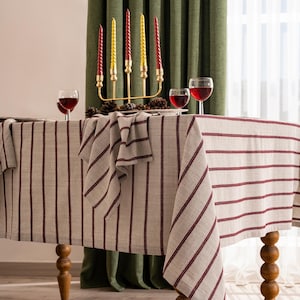 Könnte beinhalten: Ein festlich gedeckter Esstisch. Der Tisch ist mit einer cremefarbenen Tischdecke mit vertikalen bordeauxfarbenen Streifen bedeckt. Ein goldener Kerzenständer hält rote und gelbe Kerzen. Weingläser und Tannenzapfen vervollständigen die Szene.