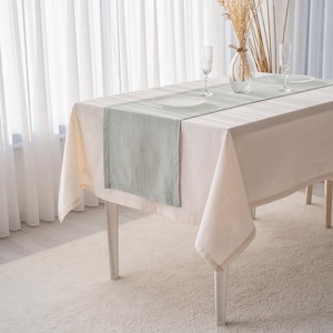 May include: A rectangular dining table set for a meal. The table is covered with a cream-colored tablecloth and a light green table runner. Two clear wine glasses and plates are set on the table. A vase with dried flowers sits in the center.