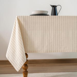 Könnte beinhalten: Eine cremefarbene Tischdecke mit vertikalen schwarzen Streifen bedeckt einen Holztisch. Eine schwarze Kanne und ein Teller mit schwarzem Rand stehen darauf. Der Tisch hat ein dekoratives, gedrechseltes Bein.