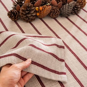 Puede incluir: Una servilleta de lino plegada con rayas beige y burdeos. La servilleta se sostiene en una mano, con un fondo de un mantel a rayas a juego y pi&ntilde;as. Las rayas est&aacute;n espaciadas uniformemente y son diagonales.