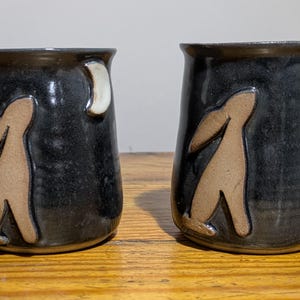 May include: Two ceramic mugs with a dark, textured glaze. Each mug features a tan figure and a white crescent moon design. The mugs are displayed on a wooden surface.