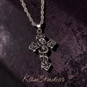 May include: Silver-toned cross pendant necklace featuring a skull and snake design. The cross has ornate detailing and hangs from a rope chain. The image is set against a dark background, with the text "KauStudios" in the lower right corner.