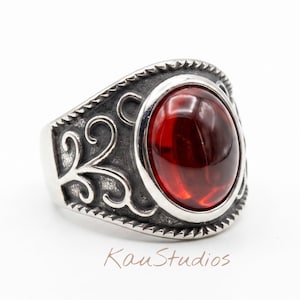 May include: A silver ring featuring a large, oval, deep red cabochon stone set in the center. The band is crafted with oxidized silver, showcasing intricate scrollwork patterns and a rope-style border. The text KauStudios is visible below.