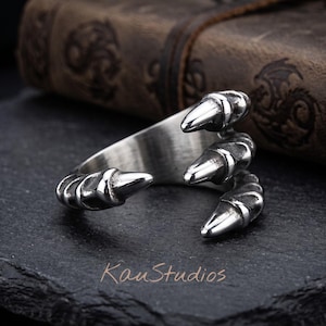 May include: A silver-toned metal ring shaped like a three-pronged claw with a polished finish. The band features a textured, segmented design that wraps around the finger. The text KauStudios is visible in the lower portion of the image.