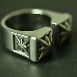 May include: A silver-toned metal ring featuring an open-ended design with two square-shaped terminals. Each terminal is decorated with a raised, multi-pointed star or leaf-like motif set against a recessed, darkened background on the band.