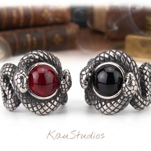 Könnte beinhalten: Zwei silberfarbene Metallringe mit einem gewundenen Schlangendesign. Ein Ring enthält einen glatten, runden, tiefroten Stein, während der andere Ring einen glatten, runden, schwarzen Stein aufweist. Der Text KauStudios steht unter den Ringen.