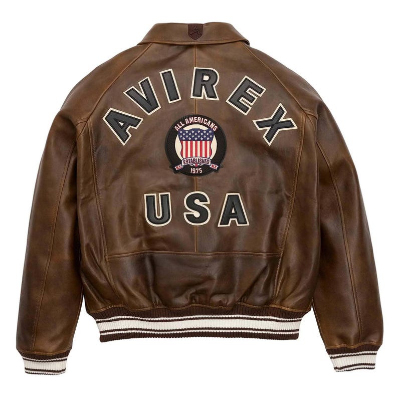 Puede incluir: Chaqueta de cuero marr&oacute;n con la palabra "AVIREX" en los hombros y "USA" en la parte inferior. Un parche circular en el centro presenta la bandera estadounidense y las palabras "ALL AMERICANS ESTABLISHED 1975". La chaqueta tiene cuello y pu&ntilde;os acanalados con rayas blancas.