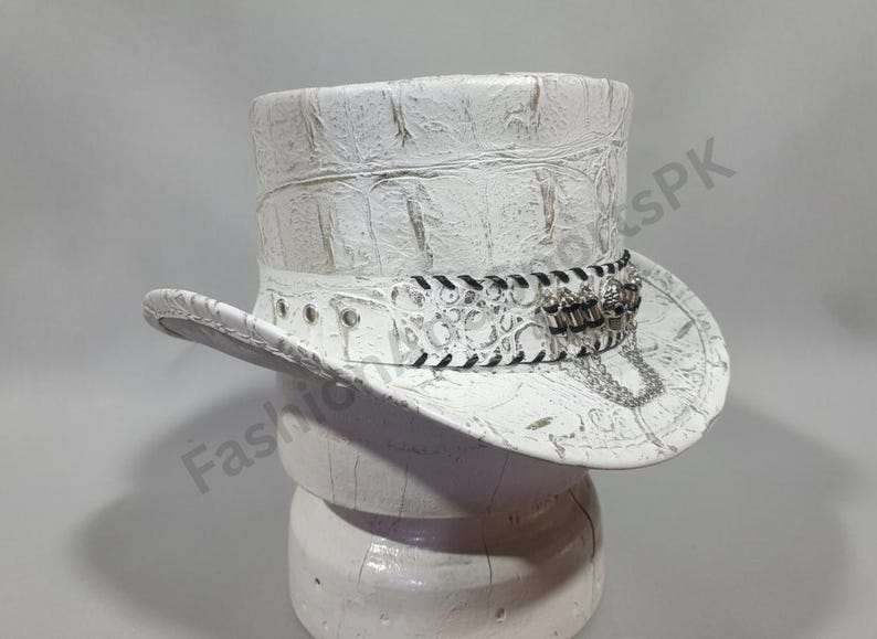 May include: White top hat with a textured, crocodile-like pattern. The hat features a decorative band with black stitching, silver-toned embellishments, and a chain. The hat is displayed on a white stand.
