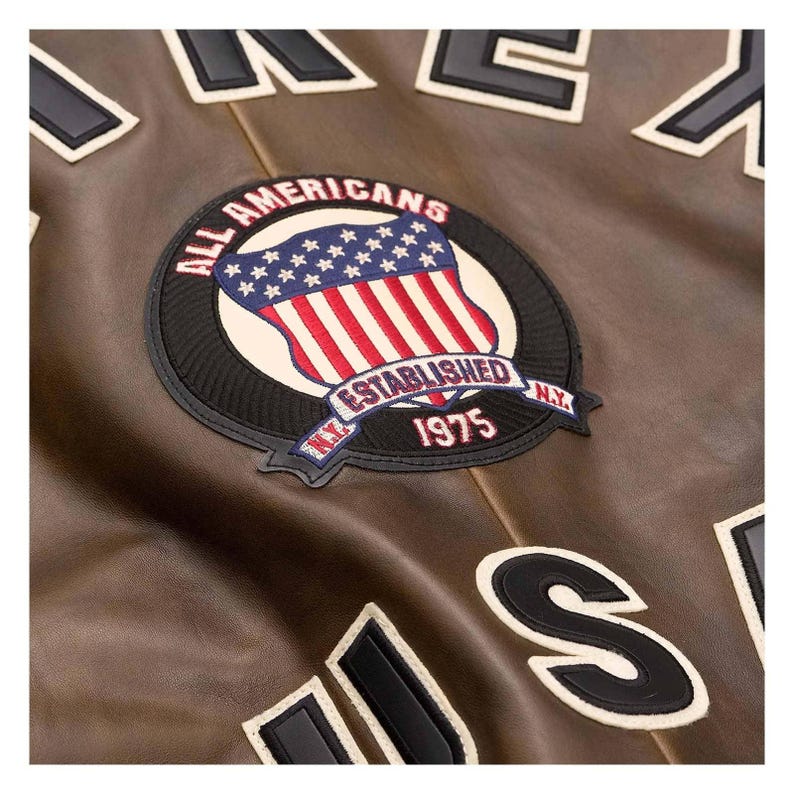 Puede incluir: Primer plano de una chaqueta de cuero marr&oacute;n con un parche que dice "ALL AMERICANS ESTABLISHED 1975 N.Y." en un dise&ntilde;o circular con una bandera de EE. UU. La chaqueta tiene letras blancas y negras.