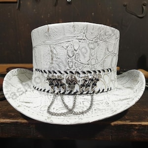 Puede incluir: Sombrero de copa blanco con un patrón texturizado similar al de un cocodrilo. El sombrero presenta una banda en blanco y negro con adornos de calaveras y dagas, y una cadena plateada. Un accesorio único.