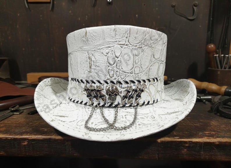 May include: White top hat with a textured, crocodile-like pattern. The hat features a black and white band with skull and dagger embellishments, and a silver chain. A unique accessory.
