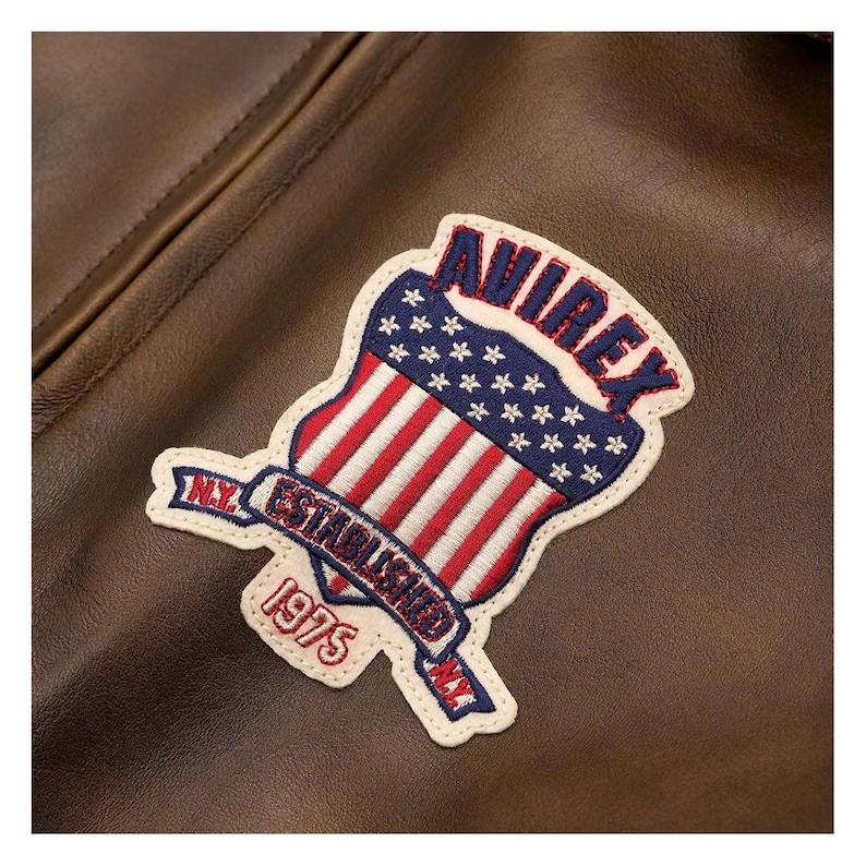 Puede incluir: Primer plano de una chaqueta de cuero marr&oacute;n con un parche. El parche tiene forma de escudo con rayas rojas y blancas y estrellas azules, con la palabra "AVIREX" en azul. El parche tambi&eacute;n tiene las palabras "N.Y. ESTABLISHED 1975 N.Y."