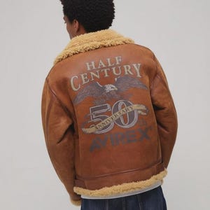 Könnte beinhalten: Braune Lederjacke mit beigem Lammfellkragen und Futter. Auf dem Rücken befindet sich eine Grafik mit einem Adler, den Worten "HALF CENTURY", "50 ANNIVERSARY" und dem Markennamen "AVIREX".