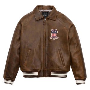 Puede incluir: Chaqueta de cuero marr&oacute;n con cuello y cierre de cremallera. La chaqueta presenta un parche en el pecho con un dise&ntilde;o de bandera americana y la marca "AVIREX". Los pu&ntilde;os y el dobladillo tienen un dise&ntilde;o a rayas en blanco y marr&oacute;n.