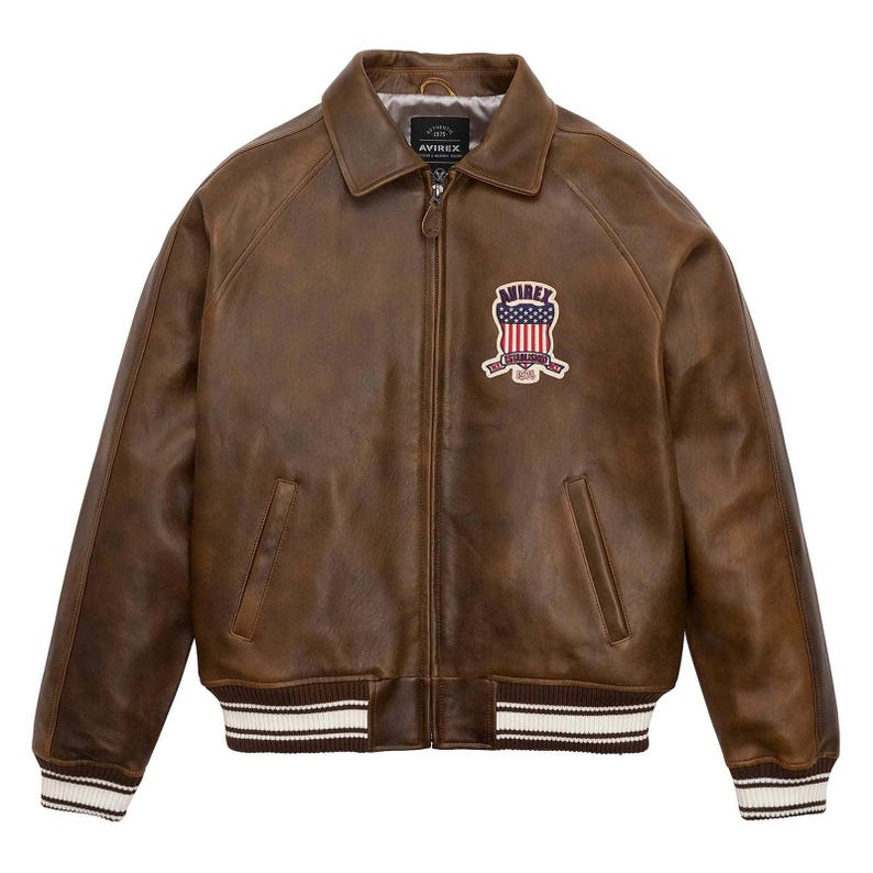 Puede incluir: Chaqueta de cuero marr&oacute;n con cuello y cierre de cremallera. La chaqueta presenta un parche en el pecho con un dise&ntilde;o de bandera americana y la marca "AVIREX". Los pu&ntilde;os y el dobladillo tienen un dise&ntilde;o a rayas en blanco y marr&oacute;n.