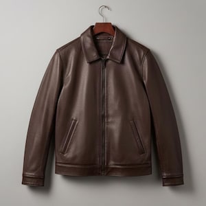 May include: A brown leather jacket with a collared neckline and a front zipper closure. The jacket has two front pockets and long sleeves. It is hanging on a wooden hanger against a neutral gray background.