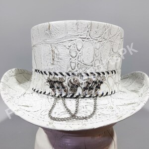 May include: White top hat with a textured, faux-leather finish. The hat features a decorative band with a skull and chain accents. The brim is wide and the hat has a classic top hat shape.