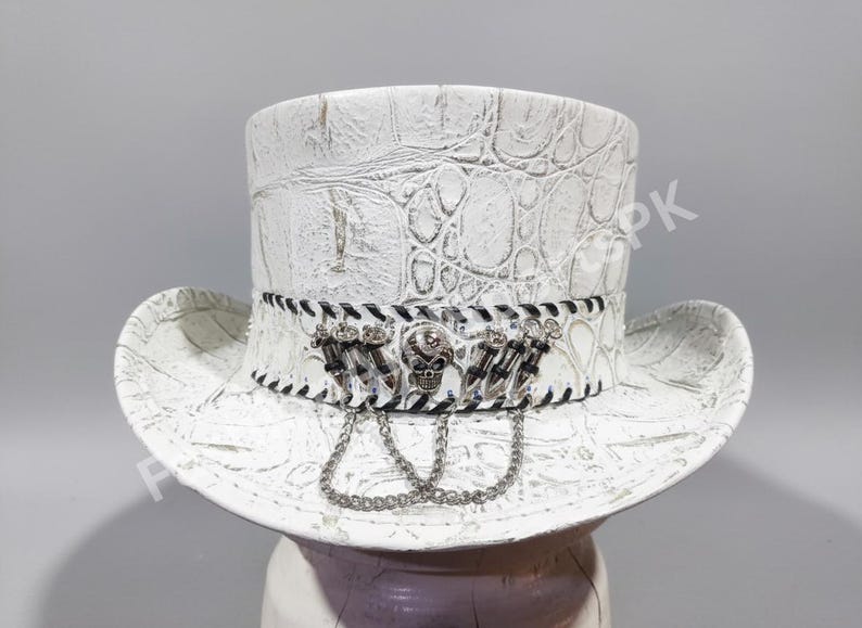 May include: White top hat with a textured, faux-leather finish. The hat features a decorative band with a skull and chain accents. The brim is wide and the hat has a classic top hat shape.
