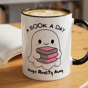 May include: White ceramic mug with a black handle and rim, featuring a cartoon ghost holding pink books. The mug has the text "A Book A Day Keeps Reality Away". The mug is filled with a coffee drink.