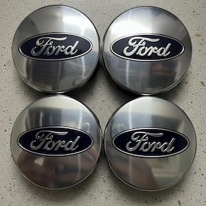 Puede incluir: Cuatro tapacubos Ford plateados con acabado metálico cepillado. Cada tapacubo presenta el logotipo de Ford en un óvalo azul oscuro. Los tapacubos están dispuestos en forma cuadrada.