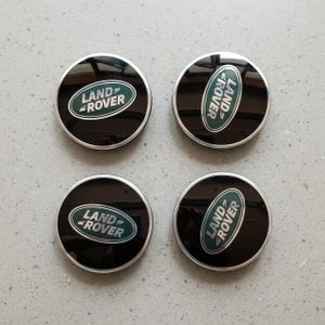 May include: Four black Land Rover wheel center caps with a silver trim and a green and white logo. The Land Rover logo is centered on each cap, with the text "LAND" above "ROVER".
