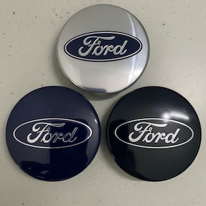 May include: Three Ford hubcaps in silver, navy blue, and black. Each circular hubcap displays the Ford logo in a contrasting colour. The hubcaps have a glossy finish and appear to be new.