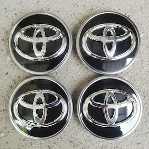 May include: Four black and silver Toyota wheel center caps. Each cap features the Toyota logo in silver against a black background, surrounded by a silver ring. The caps are circular and appear to be new.