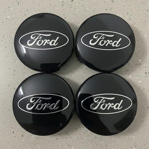 Puede incluir: Cuatro tapacubos negros Ford con acabado brillante. Cada tapacubo presenta el logotipo de Ford en un diseño ovalado plateado. Los tapacubos están dispuestos en forma cuadrada, mostrando su forma circular y superficie reflectante.