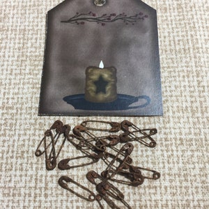 May include: A brown tag with a candle illustration and a jute string, accompanied by several rusty safety pins. The candle has a star design and a lit flame. The tag has a branch with red berries.
