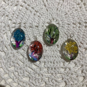 May include: Four oval-shaped glass pendants with silver chains. Each pendant contains a pressed flower arrangement depicting a tree in a different color: blue, red, green, and yellow.