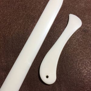 May include: Two white bone folders for crafting. One is long and straight with a pointed end, and the other is curved with a rounded end and a small hole. These tools are used for scoring and folding paper.