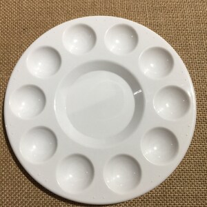 May include: A round, white ceramic paint palette with 13 wells. The palette has a central mixing area and is designed for holding and mixing paints. The palette is sitting on a brown, textured surface.