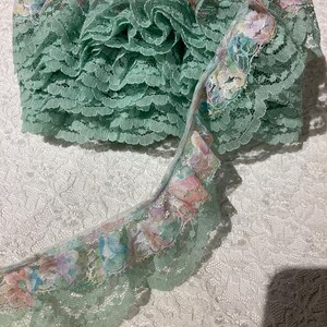 May include: A roll of mint green lace with a ruffled edge. A section of the lace is unrolled, revealing a floral pattern in pastel colors. The lace is likely used for sewing, crafting, or adding a decorative touch to garments.