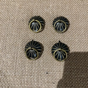 May include: Four antique-style cabinet pulls. Each pull features a dark, ridged, circular base with a gold-colored ring handle. The pulls are arranged on a textured, tan-colored surface.