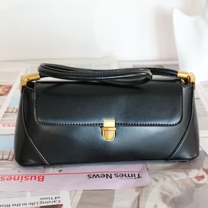 Puede incluir: Un bolso negro de forma rectangular con un cierre dorado. El bolso tiene un asa corta y detalles dorados en los laterales. El bolso está sobre un periódico.