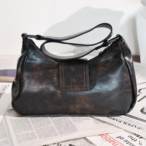 May include: A dark brown shoulder bag with a distressed leather appearance. The bag features a curved shape, a single shoulder strap, and a rectangular flap with a buckle closure. The bag is set against a background of newspaper print.