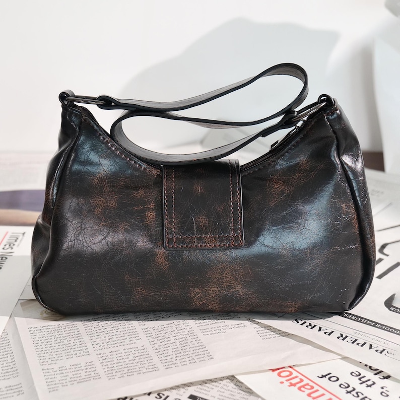 May include: A dark brown shoulder bag with a distressed leather appearance. The bag features a curved shape, a single shoulder strap, and a rectangular flap with a buckle closure. The bag is set against a background of newspaper print.