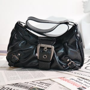 May include: A glossy black handbag with a silver buckle closure and two smaller buckles. The bag has a curved handle and sits on a newspaper background.