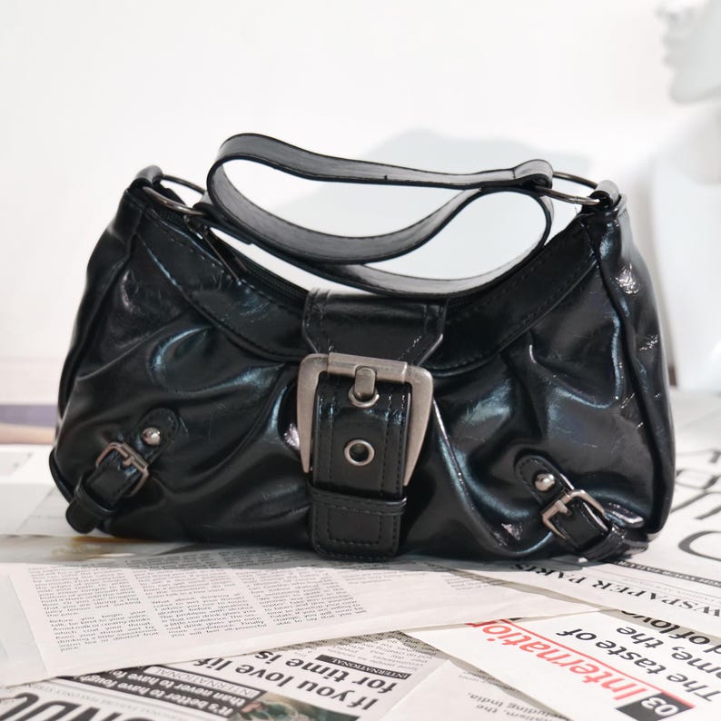 May include: A glossy black handbag with a silver buckle closure and two smaller buckles. The bag has a curved handle and sits on a newspaper background.