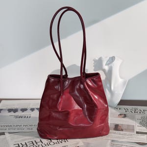 Puede incluir: Un bolso tote de cuero burdeos con asas largas y finas. El bolso tiene un acabado brillante y un aspecto ligeramente arrugado. El fondo incluye un busto blanco y recortes de periódico.